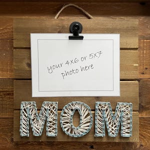 May include: Wooden photo display with the word "MOM" crafted with string art. A black clip holds a white card with the text "your 4x6 or 5x7 photo here". The background is made of wooden planks, and a jute rope is attached for hanging.