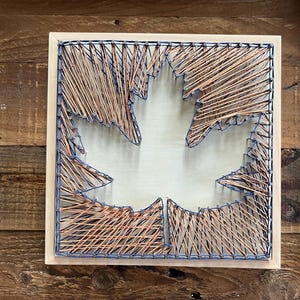 Puede incluir: Arte mural cuadrado de madera con un diseño de hoja de arce. La hoja está creada con hilo color cobre, delineada con hilo azul, y colocada sobre un fondo claro. La obra de arte está enmarcada en madera natural.