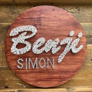 Può includere: Un cartello rotondo in legno tinto con i nomi "Benji" e "Simon" realizzati con l'arte della stringa. Il cartello è di un profondo color marrone rossiccio, e lo spago è bianco.