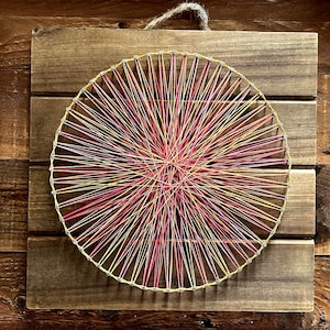 May include: A handmade string art piece featuring a circular design with red, yellow, and pink threads. The artwork is mounted on a square wooden board with a natural finish, and a jute loop for hanging.
