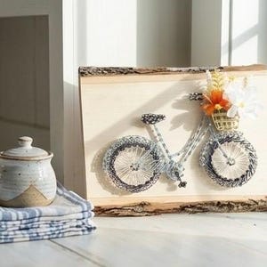 May include: A string art bicycle with a basket of flowers is mounted on a wooden plaque. The bicycle is made with silver and black thread. A small ceramic jar with a lid sits on a stack of blue and white striped napkins.