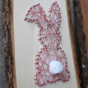 Op de afbeelding: Een houten paashaas string art stuk. De haas is omrand met spijkers en rode en witte draad, met een pluizige witte katoenen staart. Het kunstwerk is gemonteerd op een lichtgekleurde houten plank met een natuurlijke schorsrand.