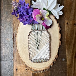 May include: A string art mason jar design created with white thread and copper-colored nails on a light-colored oval wood slice. The jar is filled with artificial purple lilac sprigs, a large white flower, and a purple and yellow pansy blossom.