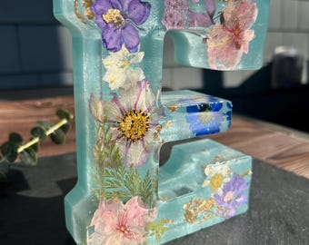 Könnte beinhalten: Ein türkisfarbener Resin-Buchstabe "E" mit eingebetteten getrockneten Blumen in Lila-, Rosa-, Weißtönen und Goldblatt-Akzenten. Das florale Design ist im gesamten Buchstaben sichtbar und schafft ein einzigartiges Dekorationsobjekt.