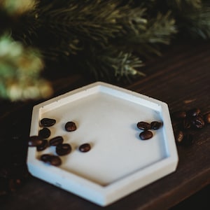 Pode incluir: Uma bandeja hexagonal de cerâmica branca com uma borda elevada, cheia de grãos de café. A bandeja está sobre uma superfície de madeira escura.