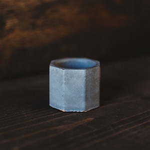 May include: A small, hexagonal concrete planter in a light gray color. The planter is sitting on a dark wood surface.