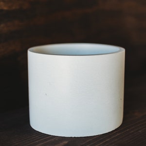 Puede incluir: Un macetero cilíndrico de cerámica blanca, perfecto para exhibir plantas pequeñas o suculentas. El macetero tiene un acabado liso y mate y un fondo ligeramente redondeado.