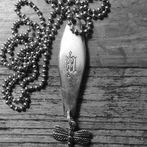 May include: A silver necklace with a charm in the shape of a dragonfly. The charm is attached to a silver spoon with an engraved letter 'H'. The necklace is made of a silver chain.