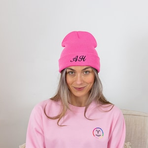 Op de afbeelding: Een roze beanie met de initialen "A.H." geborduurd in het zwart. De beanie wordt gedragen door iemand die een roze sweatshirt draagt met een regenboogkleurig sterrenbeeld voor Ram.
