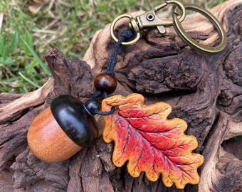 May include: A handcrafted keychain featuring a wooden acorn and a colorful leaf pendant. The acorn has a dark cap and a light brown body. The leaf is painted with shades of red, orange, and yellow, attached to a bronze-colored clasp.