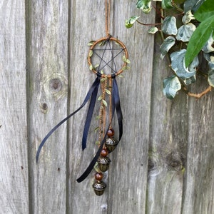 May include: A brown and black dreamcatcher with a pentagram design and green leaves. It has black ribbons and three gold bells hanging from it.