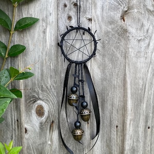 May include: A black dreamcatcher featuring a pentagram design, black ribbons, and acorn-shaped charms. It hangs against a weathered wooden fence. The background includes green foliage.