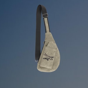 May include: A light gray sling bag with a black strap and a small front pocket. The pocket features a black embroidered airplane design. The bag is made of a textured material and has a zipper closure.