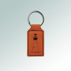 May include: A brown leather keychain with a silver ring. The keychain features a black illustration of a lighthouse. The lighthouse is on a rectangular tag with a stitched border.