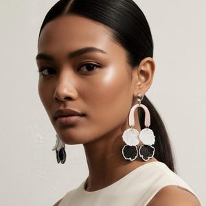 Puede incluir: Pendientes llamativos con un arco rosa pálido y tres racimos de flores con cuentas en blanco y negro. Los pendientes cuelgan, con una pequeña flor en blanco y negro en la oreja izquierda. La persona lleva un vestido crema sin mangas.