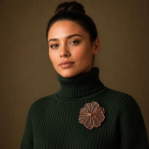 Puede incluir: Un broche de flores calado de color cobre está prendido a un jersey de cuello alto verde oscuro. El broche tiene un diseño detallado y en capas. La mujer de la imagen tiene el pelo oscuro y mira a la cámara.