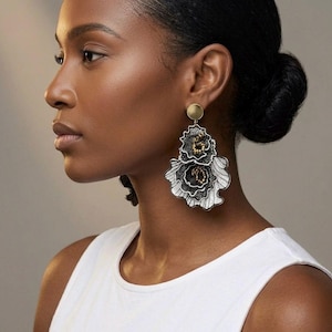 Puede incluir: Elegantes pendientes florales con un perno dorado y detalles intrincados en negro, plateado y dorado. Los pendientes presentan un diseño en capas que se asemeja a flores estilizadas.