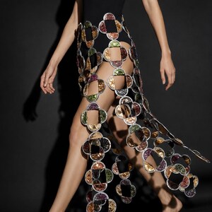 Puede incluir: Un body negro de un solo hombro combinado con una falda decorativa construida con discos metálicos circulares interconectados en tonos verde, morado, cobre y plata, con un diseño calado que crea un patrón repetitivo en forma de trébol.