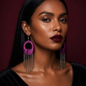 Puede incluir: Una mujer lleva pendientes colgantes grandes. Los pendientes presentan un diseño circular de color magenta con flecos de cadena plateada. La mujer tiene el pelo oscuro, lápiz labial oscuro y lleva una blusa negra. El fondo es de color burdeos oscuro.