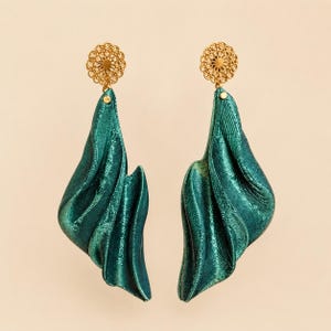 Puede incluir: Un par de pendientes colgantes con postes circulares de filigrana en tono dorado unidos a largos drapeados de color verde esmeralda oscuro, fluidos y con una textura metálica ondulada. Los pendientes tienen una forma escultural.