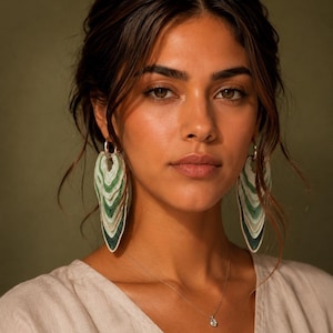 Puede incluir: Un par de pendientes colgantes con elementos superpuestos en forma de hoja en tonos verde bosque, salvia y crema. Los pendientes cuelgan de aros plateados sencillos y se llevan con un collar de cadena fina de plata con un pequeño colgante.