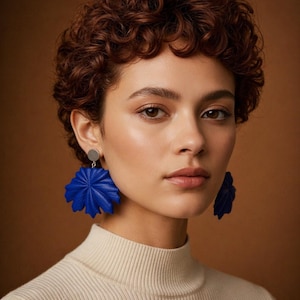 Puede incluir: Una mujer con el pelo corto y rizado de color castaño rojizo lleva pendientes grandes de color azul brillante. Los pendientes tienen un diseño en capas, similar a un pétalo, y están suspendidos de pequeños tachuelas plateadas. Lleva un jersey de cuello alto color crema. El fondo es de color marrón cálido.