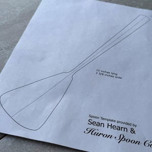 Op de afbeelding: Een zwart-witte lijntekening van een lepel sjabloon op een wit vel papier. De sjabloon is 11 inch lang en 2 3/8 inch breed. De tekst "Spoon Template provided by Sean Hearn & Huron Spoon Co" is onder de sjabloon afgedrukt.