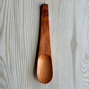May include: A handcrafted wooden spoon with a long handle and a rounded bowl. The spoon is made of light brown wood with darker brown grain patterns. The handle is approximately 12 inches long.