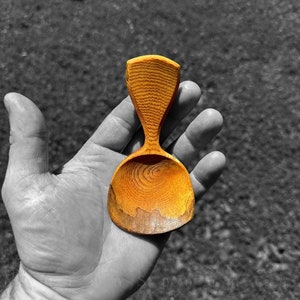 May include: A small, hand-carved wooden spoon with a rounded bowl and a unique, slightly pointed handle. The spoon is made of light wood with a natural, warm color.