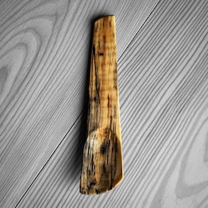 May include: A handmade wooden spoon with a natural, organic aesthetic. The spoon features a light brown color with dark brown and black grain patterns. The spoon's design includes a rounded bowl and a long, flat handle. The spoon is likely used for cooking or serving.