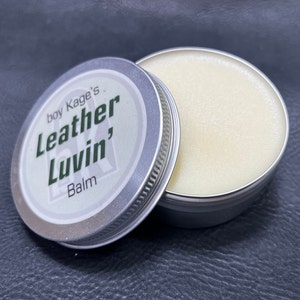 Puede incluir: Una lata de plata con una etiqueta blanca que dice "boy Kage's Leather Luvin' Balm". La lata está abierta y el bálsamo en el interior es de color amarillo claro.