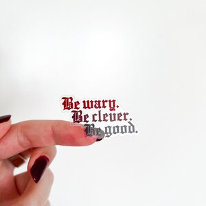 Può includere: Un adesivo trasparente con il testo "Be wary. Be clever. Be good." in un font gotico. Il testo è in rosso e nero.