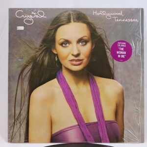 May include: A vinyl record album cover featuring a woman with long brown hair wearing a purple top and a purple scarf. The album title is "Crystal" and the text "Hollywood, Tennessee" is printed above the woman's head. The album features the single "The Woman In Me".