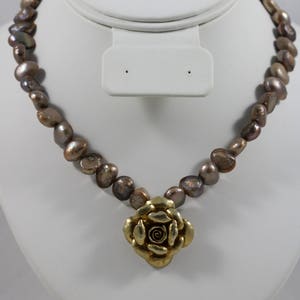 May include: A brown pearl necklace with a gold rose pendant. The necklace is displayed on a white mannequin bust.
