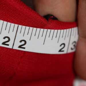 May include: Close-up of a white measuring tape with black markings. The tape reads "2 2 2 3". The tape is lying on top of a red fabric.