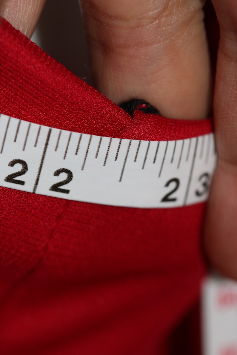 May include: Close-up of a white measuring tape with black markings. The tape reads "2 2 2 3". The tape is lying on top of a red fabric.
