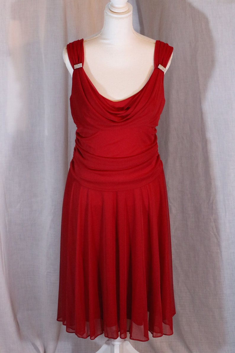 May include: A red sleeveless dress with a cowl neck and a flowing skirt. The dress has a jeweled embellishment on each shoulder strap.
