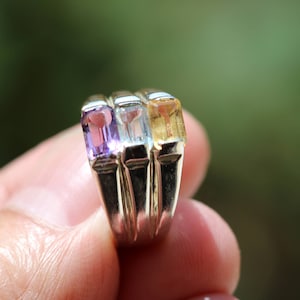 May include: A silver ring with three rectangular gemstones. The gemstones are purple, light blue, and yellow.