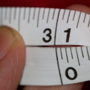 May include: A white measuring tape with black markings shows the numbers 31 and 0. The tape is being held by a finger.