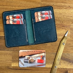 Peut inclure: Portefeuille en cuir bleu canard ouvert, avec des coutures blanches, contenant des cartes. Une carte de visite avec le texte "Jimmy Afer Cuir & Artisanat" et un couteau en laiton sont posés sur une surface en bois.