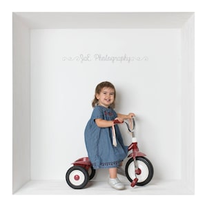 May include: A young girl in a blue denim dress rides a red tricycle with white wheels in a white room. The text "Jae Photography" is written on the wall.