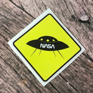 May include: A yellow warning sign with a black silhouette of a UFO with the word "NASA" printed on it.