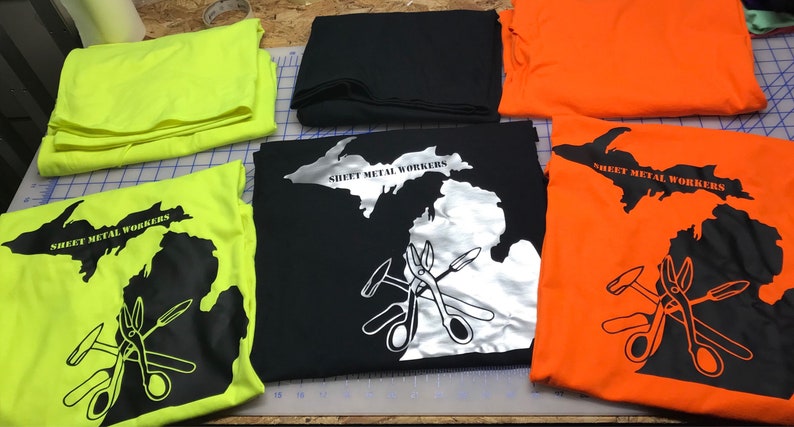 May include: Four t-shirts with a black and white outline of the state of Michigan with the text "Sheet Metal Workers" and a graphic of tools. The shirts are in the colors yellow, black, orange, and white.