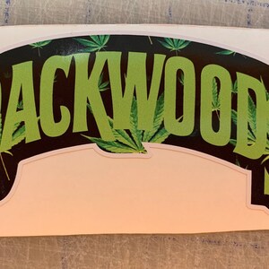 Può includere: Un adesivo con la scritta "BACKWOODS" in grandi lettere verde brillante. Le lettere sono bordate di nero e poste su uno sfondo nero con foglie di cannabis verdi. L'adesivo ha un bordo bianco.