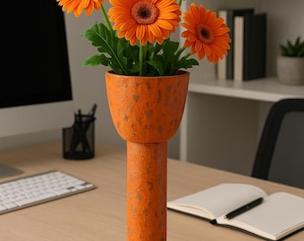 May include: A tall, orange vase with a textured finish holds a bouquet of vibrant orange gerbera daisies with dark brown centers and green leaves. The vase sits on a wooden desk, with a computer and open notebook in the background.