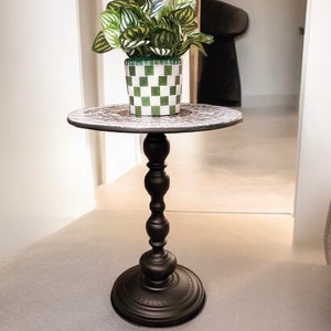 Pode incluir: Uma mesa auxiliar preta com tampo redondo de mosaico e base decorativa. Um vaso verde e branco xadrez com uma planta verde está sobre a mesa.