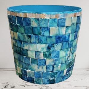 Puede incluir: Un jarrón decorativo de mosaico con interior y borde azules. El exterior está cubierto con un patrón de mosaico de baldosas cuadradas azules, turquesas y blancas. El jarrón tiene forma cilíndrica.