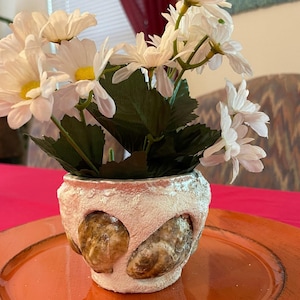 Op de afbeelding: Een witte keramische pot met een getextureerd oppervlak en een ontwerp van schelpen. De pot is gevuld met witte bloemen.