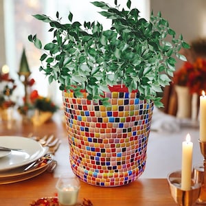 Puede incluir: Una vibrante maceta de mosaico llena de exuberante follaje verde. La maceta es cilíndrica, adornada con un colorido patrón de mosaico de pequeñas baldosas cuadradas en varios tonos. Las hojas de la planta caen sobre el borde, creando un contraste llamativo.
