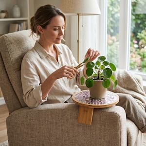 May include: A round wooden armrest tray with a colourful mosaic tile inlay, featuring a bamboo base that clips onto a chair arm. A small green Pilea plant in a beige pot sits on the tray, while gold-toned scissors are used to trim the leaves.